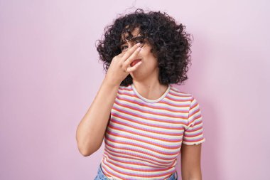 Young middle east woman standing over pink background smelling something stinky and disgusting, intolerable smell, holding breath with fingers on nose. bad smell 
