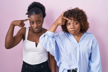 Two african women standing over pink background pointing unhappy to pimple on forehead, ugly infection of blackhead. acne and skin problem  clipart