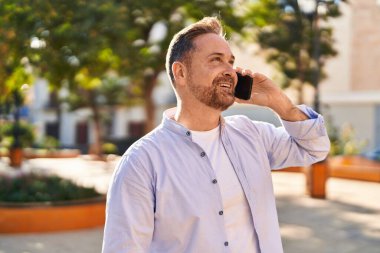 Genç beyaz adam gülümsüyor. Parktaki akıllı telefondan konuşuyor.