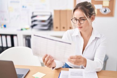 Genç sarışın iş kadını ofiste dizüstü bilgisayar okuyarak çalışıyor.