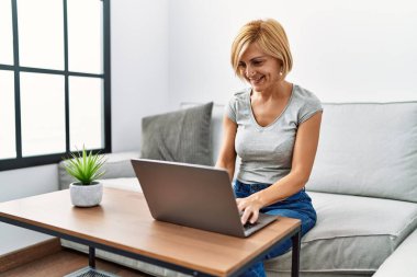 Orta yaşlı sarışın kadın evde dizüstü bilgisayar kullanarak gülümsüyor.