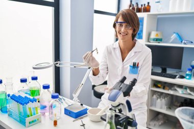 Young beautiful hispanic woman scientist smiling confident holding plant with tweezer at laboratory