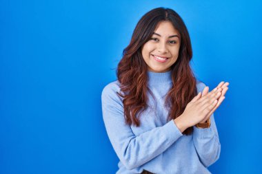 Hispanic young woman standing over blue background clapping and applauding happy and joyful, smiling proud hands together 