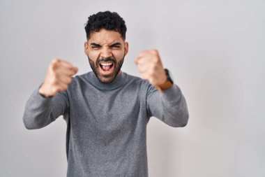 Hispanic man with beard standing over white background angry and mad raising fists frustrated and furious while shouting with anger. rage and aggressive concept. 