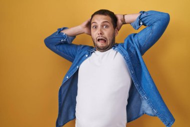 Hispanic man standing over yellow background crazy and scared with hands on head, afraid and surprised of shock with open mouth 