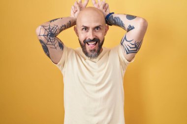 Hispanic man with tattoos standing over yellow background posing funny and crazy with fingers on head as bunny ears, smiling cheerful 