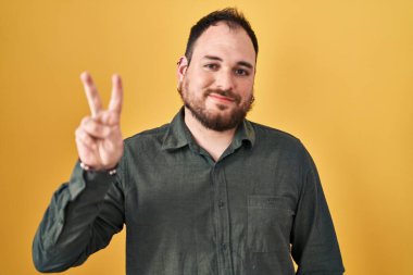 Plus size hispanic man with beard standing over yellow background showing and pointing up with fingers number two while smiling confident and happy. 