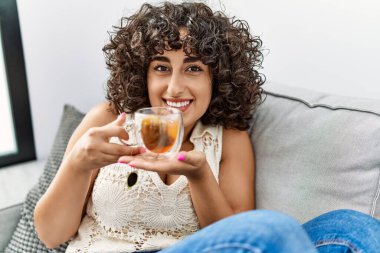 Orta Doğulu genç bir kadın gülümseyerek evde çay içiyor.