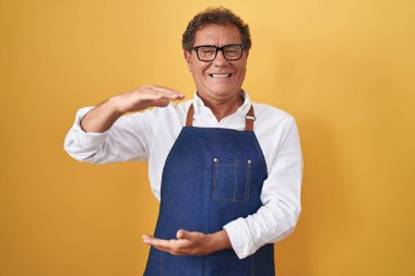 Middle age hispanic man wearing professional cook apron gesturing with hands showing big and large size sign, measure symbol. smiling looking at the camera. measuring concept. 