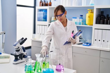 Middle age woman wearing scientist uniform holding clipboard at laboratory