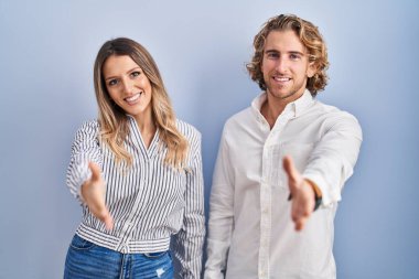 Young couple standing over blue background smiling friendly offering handshake as greeting and welcoming. successful business. 