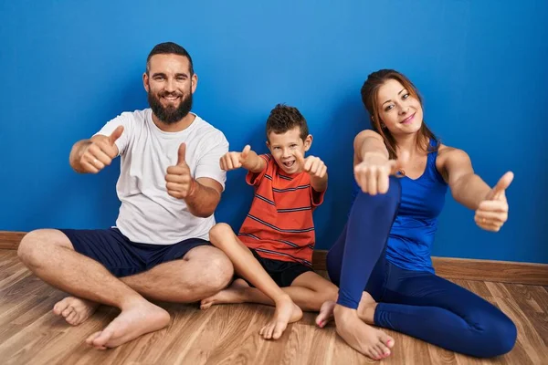 Family of three sitting on the floor at home approving doing positive gesture with hand, thumbs up smiling and happy for success. winner gesture.