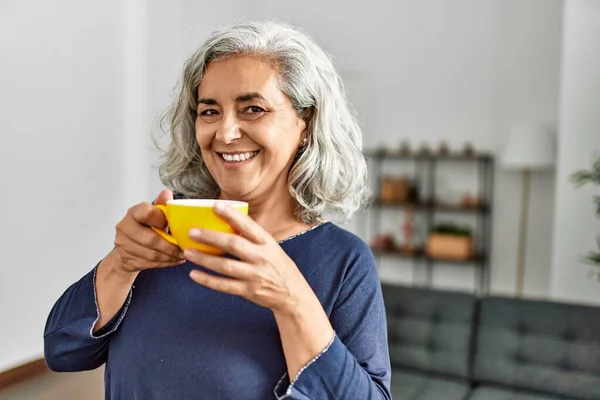 Middle age grey-haired woman drinking coffee standing at home.