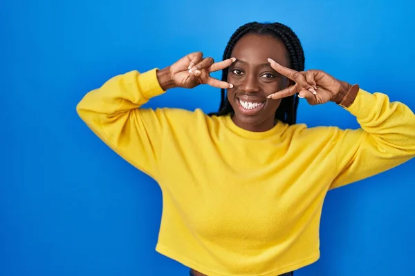 Beautiful black woman standing over blue background doing peace symbol with fingers over face, smiling cheerful showing victory