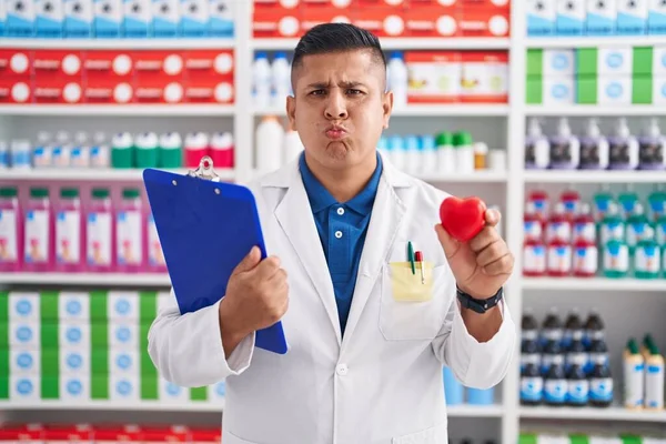 Young Hispanic Man Working Pharmacy Drugstore Holding Heart Puffing Cheeks — Stockfoto