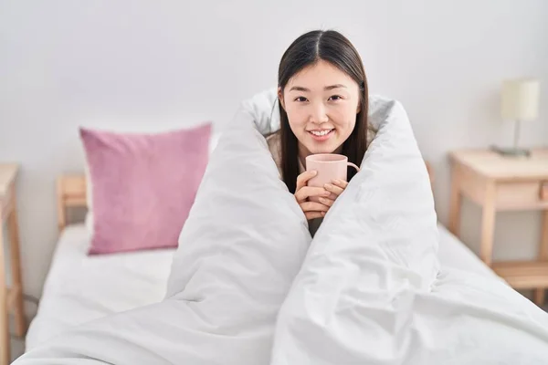 Chinese woman drinking cup of coffee covering with bedsheet at bedroom