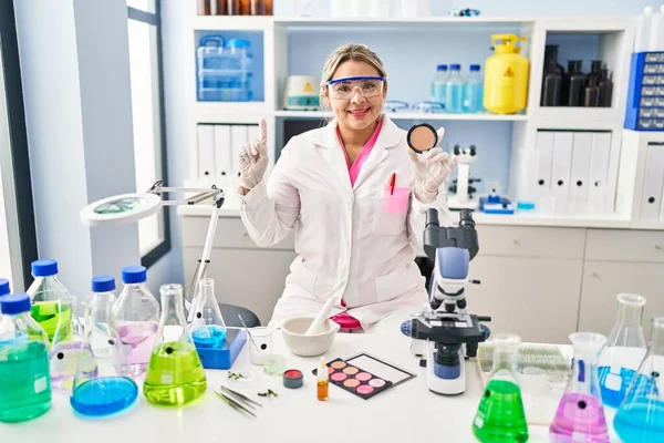 Young hispanic woman working at scientist laboratory doing make up surprised with an idea or question pointing finger with happy face, number one
