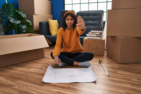 Young hispanic woman sitting on the floor at new home looking at blueprints with open hand doing stop sign with serious and confident expression, defense gesture