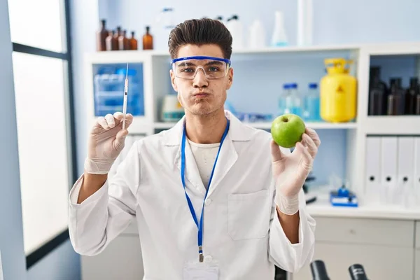 Young Hispanic Man Working Scientist Laboratory Holding Apple Puffing Cheeks —  Fotos de Stock