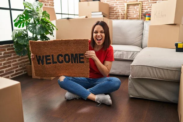 Young hispanic woman holding welcome doormat at new home angry and mad screaming frustrated and furious, shouting with anger. rage and aggressive concept.