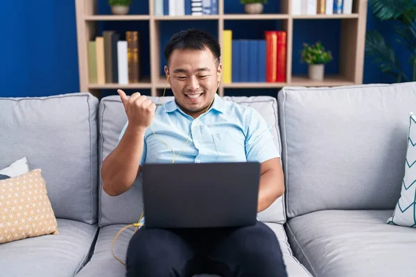Chinese young man using computer laptop sitting on the sofa pointing thumb up to the side smiling happy with open mouth