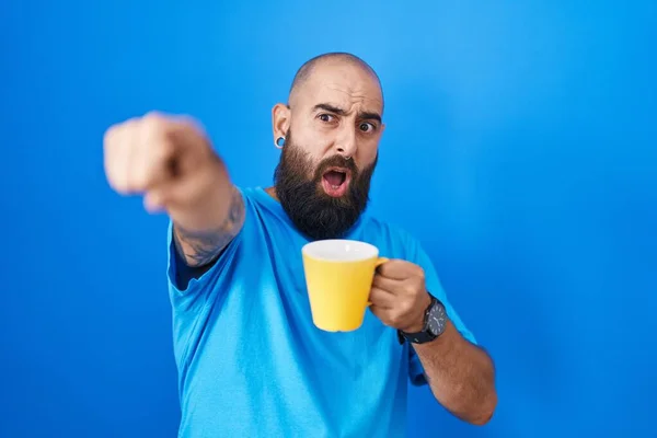 Young hispanic man with beard and tattoos drinking a cup of coffee pointing with finger surprised ahead, open mouth amazed expression, something on the front
