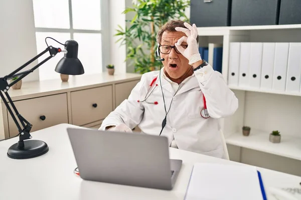 Senior doctor man working on online appointment doing ok gesture shocked with surprised face, eye looking through fingers. unbelieving expression.