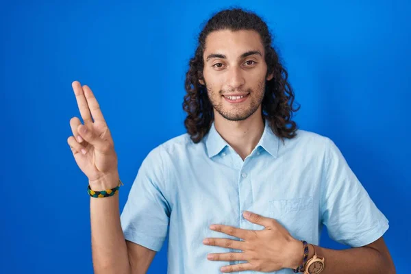 胸と指で手を差し伸べ忠誠を誓う青い背景の上に立つ若いヒスパニック系の男 — ストック写真