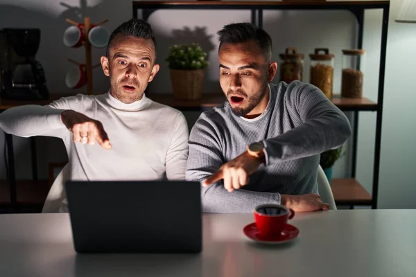 Homosexual couple using computer laptop pointing with finger surprised ahead, open mouth amazed expression, something on the front