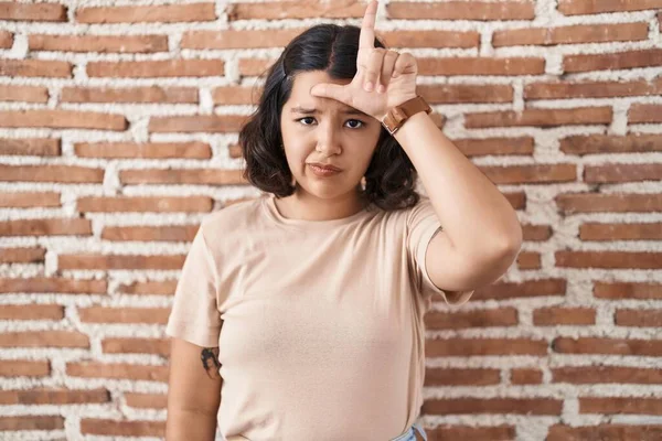 Young hispanic woman standing over bricks wall making fun of people with fingers on forehead doing loser gesture mocking and insulting.