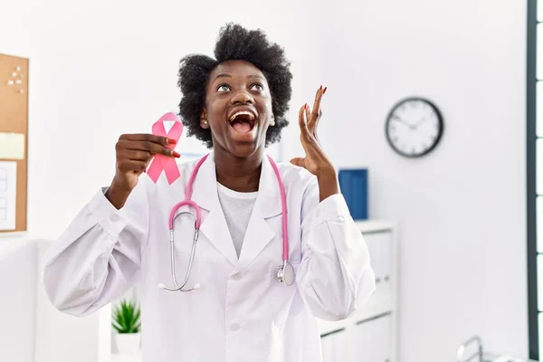 African doctor woman holding pink cancer ribbon at clinic celebrating victory with happy smile and winner expression with raised hands