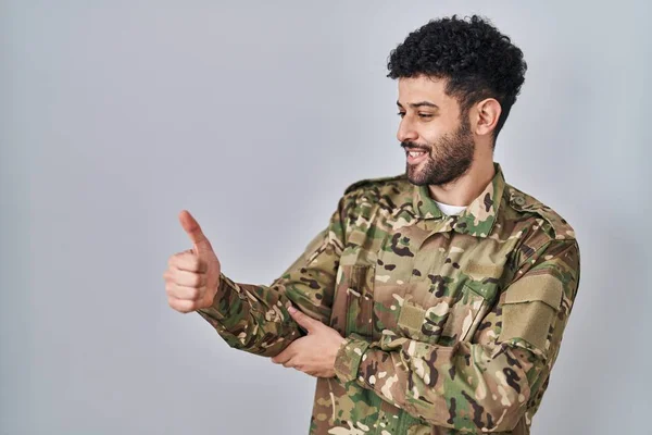 Arab man wearing camouflage army uniform looking proud, smiling doing thumbs up gesture to the side