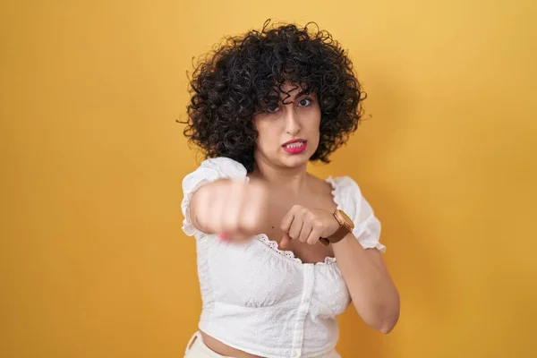 Young brunette woman with curly hair standing over yellow background punching fist to fight, aggressive and angry attack, threat and violence