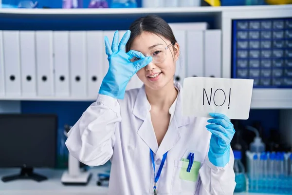 Chinese young woman working at scientist laboratory holding no banner smiling happy doing ok sign with hand on eye looking through fingers