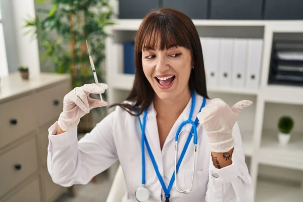 Young brunette doctor woman holding syringe pointing thumb up to the side smiling happy with open mouth