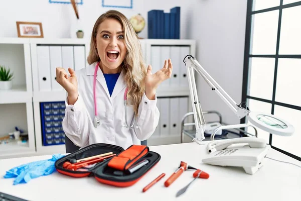 Young beautiful doctor woman with reflex hammer and medical instruments celebrating surprised and amazed for success with arms raised and open eyes. winner concept.