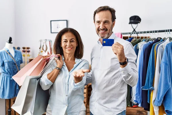 Hispanic middle age couple holding shopping bags and credit card smiling cheerful offering palm hand giving assistance and acceptance. 