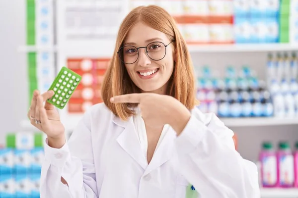 Young redhead woman working at pharmacy drugstore holding birth control pills smiling happy pointing with hand and finger