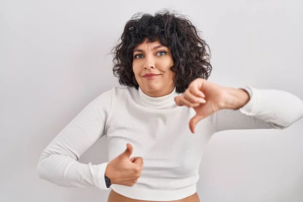 Hispanic woman with curly hair standing over isolated background doing thumbs up and down, disagreement and agreement expression. crazy conflict