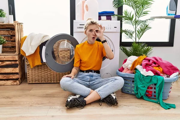 Young blonde woman doing laundry sitting by washing machine doing ok gesture shocked with surprised face, eye looking through fingers. unbelieving expression.