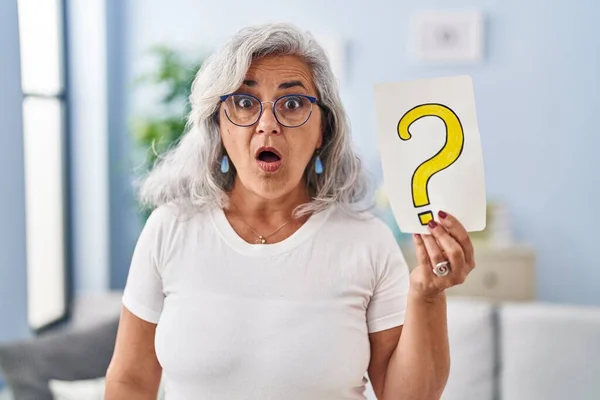 Middle age woman with grey hair holding question mark scared and amazed with open mouth for surprise, disbelief face