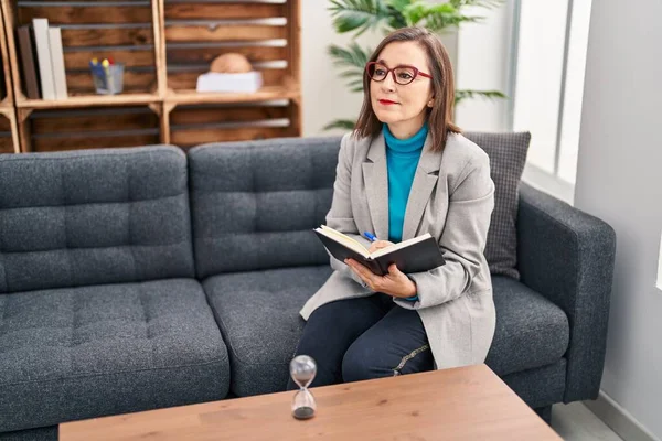 Middle age woman psychologist smiling confident reading book at psychology center