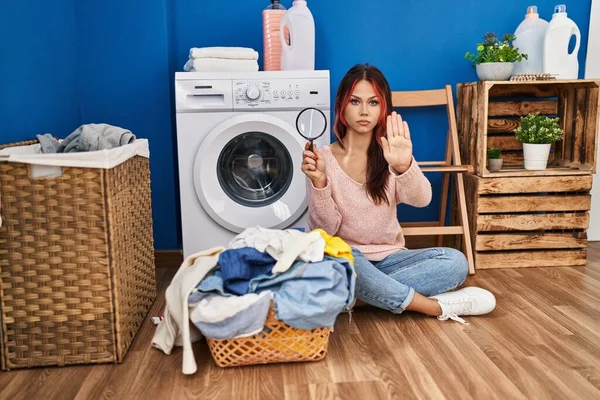 Young caucasian woman using magnifying glass glass looking for stains in dirty laundry with open hand doing stop sign with serious and confident expression, defense gesture