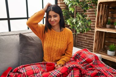 Young hispanic woman sitting on the sofa drinking a coffee at home confuse and wondering about question. uncertain with doubt, thinking with hand on head. pensive concept. 