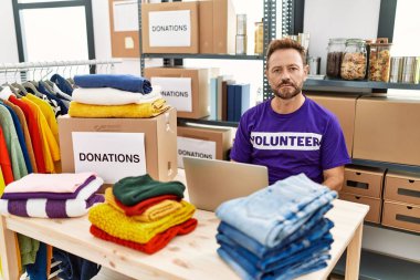 Middle age man wearing volunteer t shirt working with laptop relaxed with serious expression on face. simple and natural looking at the camera. 