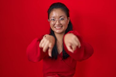 Asian young woman standing over red background pointing to you and the camera with fingers, smiling positive and cheerful 