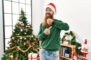 Redhead man with long beard wearing christmas hat by christmas tree in hurry pointing to watch time, impatience, looking at the camera with relaxed expression 