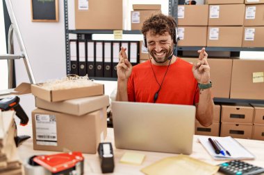 Young hispanic call center agent man working at warehouse gesturing finger crossed smiling with hope and eyes closed. luck and superstitious concept. 
