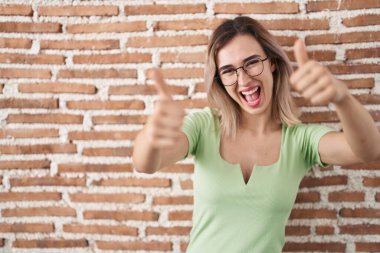 Duvarın önünde duran genç ve güzel bir kadın olumlu bir jest yapmayı onaylıyor. Başparmağıyla gülümsüyor ve başarı için seviniyor. kazanan jest. 