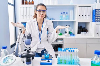 Young hispanic woman working at scientist laboratory clueless and confused expression with arms and hands raised. doubt concept. 